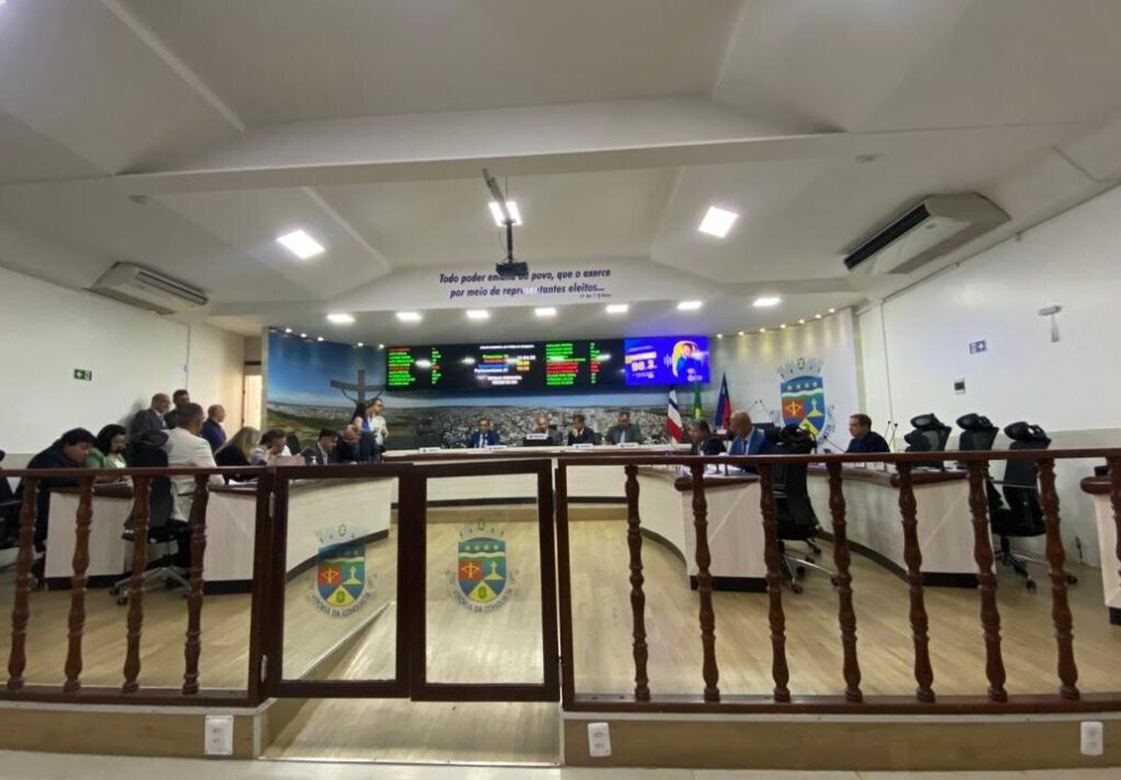 Plenário da Câmara de Vereadores com pessoas sentadas ao redor de uma grande bancada em formato de "U". Os parlamentares e assessores vestem roupas formais. Uma divisória baixa de madeira com portão de vidro separa o público do plenário.