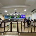 Plenário da Câmara de Vereadores com pessoas sentadas ao redor de uma grande bancada em formato de "U". Os parlamentares e assessores vestem roupas formais. Uma divisória baixa de madeira com portão de vidro separa o público do plenário.