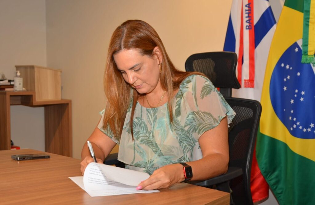 A prefeita Sheila Lemos assina um documento sobre uma mesa de madeira. Ela veste uma blusa verde clara com estampas de folhas e usa um relógio de pulso preto. Ao fundo, à direita, estão as bandeiras da Bahia e do Brasil.