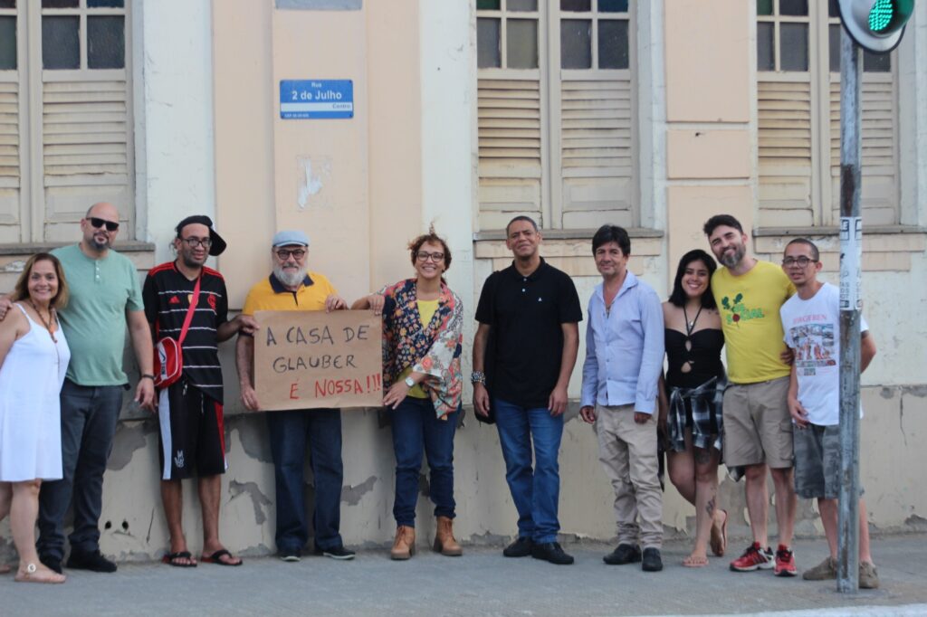 Um grupo de nove pessoas posa em uma calçada. Três pessoas no centro seguram um cartaz de papelão escrito à mão: "A CASA DE GLAUBER É NOSSA!!!". Ao fundo, a fachada de um prédio antigo exibe janelas de madeira clara e uma placa azul com o texto: "Rua 2 de Julho Centro". 