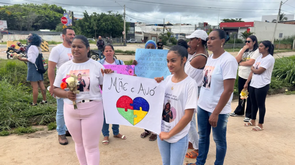 Grupo de pessoas reunidas em uma área aberta e arborizada realiza um protesto. No centro, duas mulheres seguram um cartaz branco com o texto "MÃE e AVÓ" acima de um desenho de coração em formato de quebra-cabeça colorido. Ao fundo, há cartazes manuscritos, sendo um azul com a frase: "Justiça não é vingança é o direito de quem não pode mais lutar por si mesmo".