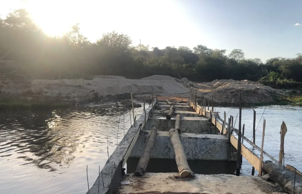 Ponte de concreto em construção sobre um rio largo. A estrutura atravessa a água, com toras de madeira e vigas metálicas aparentes sobre a base cinza. Nas margens, há montes de areia clara e vegetação densa com árvores verdes. O sol brilha intensamente ao fundo, refletindo luz amarelada na superfície da água e no céu claro.