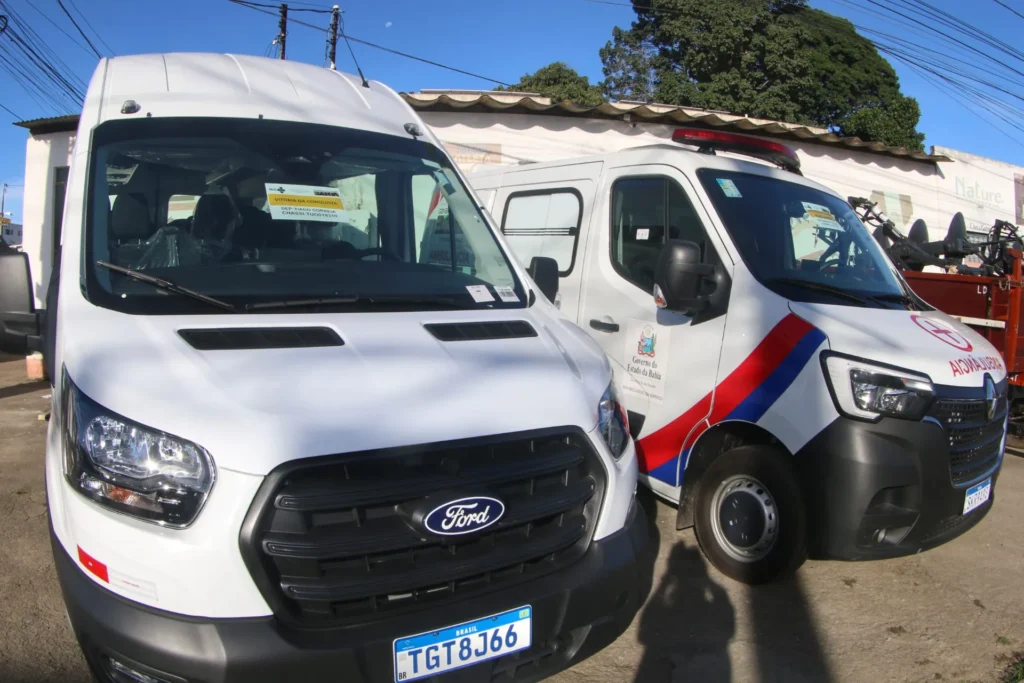 Duas ambulâncias brancas estacionadas lado a lado. No para-brisa da primeira ambulância, à esquerda, um papel indica o destino Vitória da Conquista. A segunda ambulância, à direita, possui faixas diagonais em vermelho e azul na lateral e o brasão do Governo do Estado da Bahia na porta.