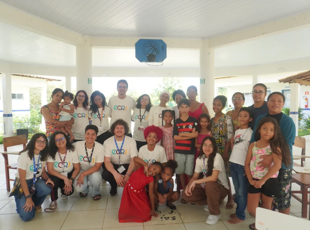 Um grupo de cerca de vinte e cinco pessoas, entre adultos e crianças, posa para uma foto em um pátio coberto. Sete adultos usam camisetas brancas com a sigla colorida ECRJ no peito e crachás no pescoço. No centro, uma menina usa um vestido vermelho vibrante e uma mulher tem cabelos curtos tingidos de vermelho. Ao fundo, o espaço é aberto, com colunas brancas, teto de madeira clara e uma tabela de basquete azul pendurada.