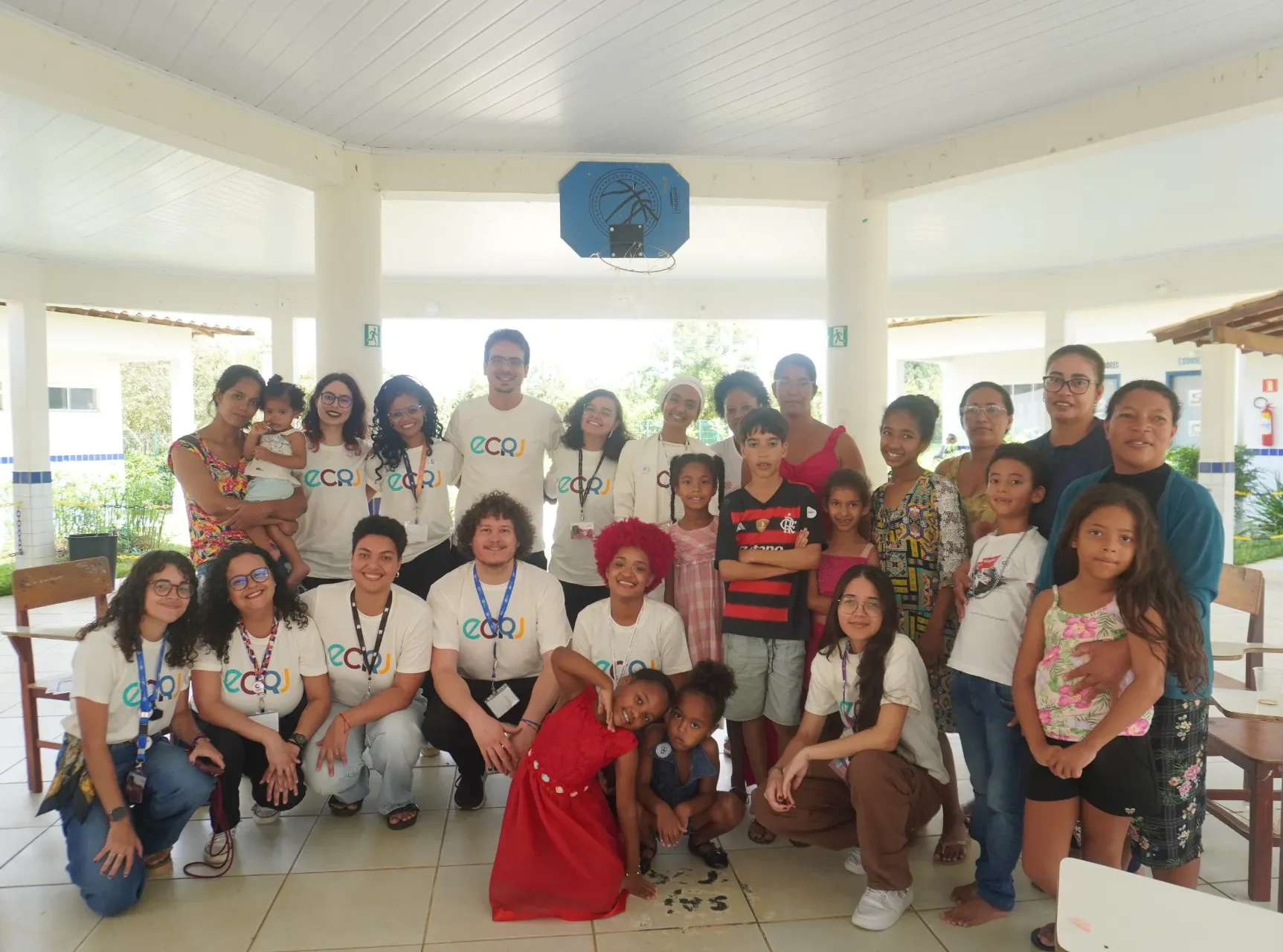Um grupo de cerca de vinte e cinco pessoas, entre adultos e crianças, posa para uma foto em um pátio coberto. Sete adultos usam camisetas brancas com a sigla colorida ECRJ no peito e crachás no pescoço. No centro, uma menina usa um vestido vermelho vibrante e uma mulher tem cabelos curtos tingidos de vermelho. Ao fundo, o espaço é aberto, com colunas brancas, teto de madeira clara e uma tabela de basquete azul pendurada.