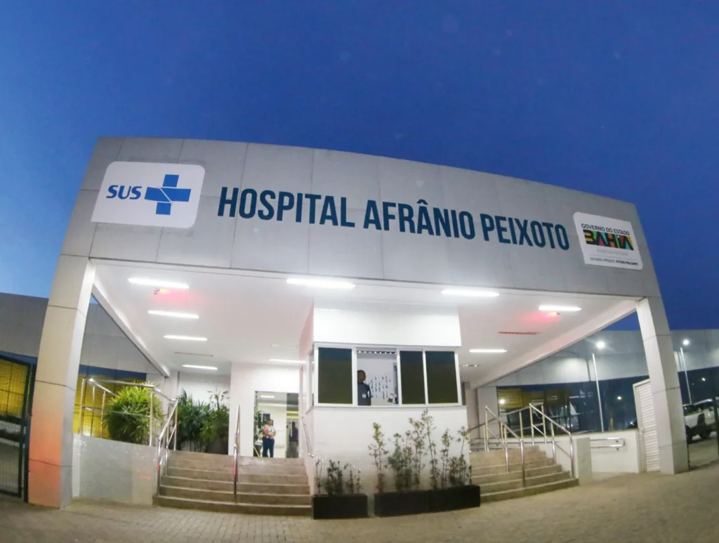 Fachada do Hospital Afrânio Peixoto sob um céu azul escuro ao entardecer. A entrada principal possui uma estrutura retangular cinza, com cobertura e escadarias. O texto na fachada diz: SUS. HOSPITAL AFRÂNIO PEIXOTO. GOVERNO DO ESTADO BAHIA. SECRETARIA DA SAÚDE.