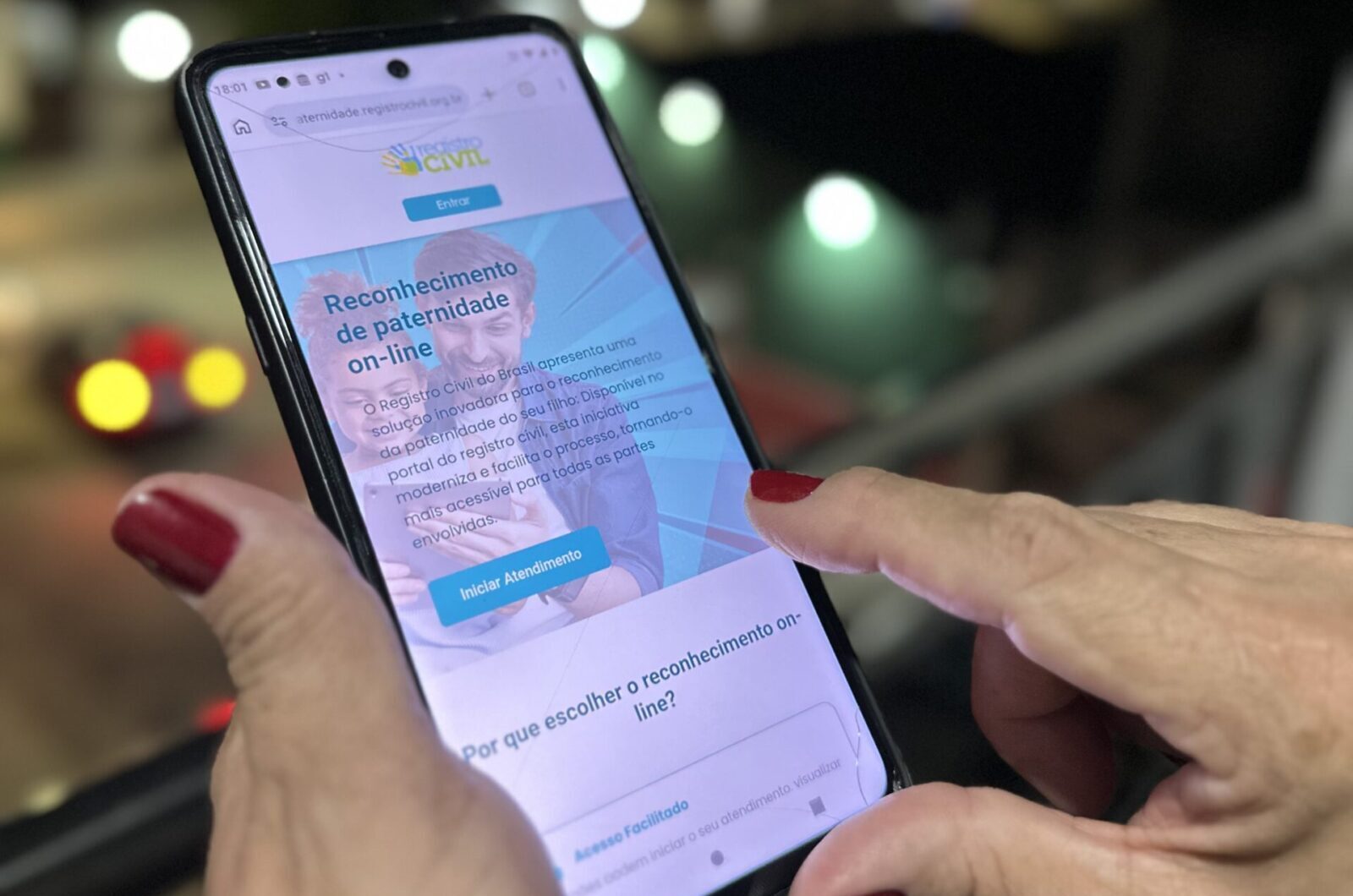Mãos de uma pessoa com unhas pintadas de vermelho seguram um celular que exibe o site do Registro Civil do Brasil sobre reconhecimento de paternidade. O aparelho está levemente inclinado, com o dedo indicador tocando a tela.