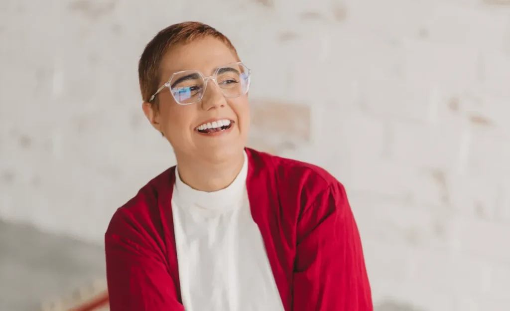Mulher de pele clara e cabelos curtos acobreados sorri e olha para a lateral. Ela usa óculos de armação transparente e quadrada, uma blusa branca e um casaco vermelho. O fundo é uma parede de tons claros e neutros com textura de tijolos aparentes.