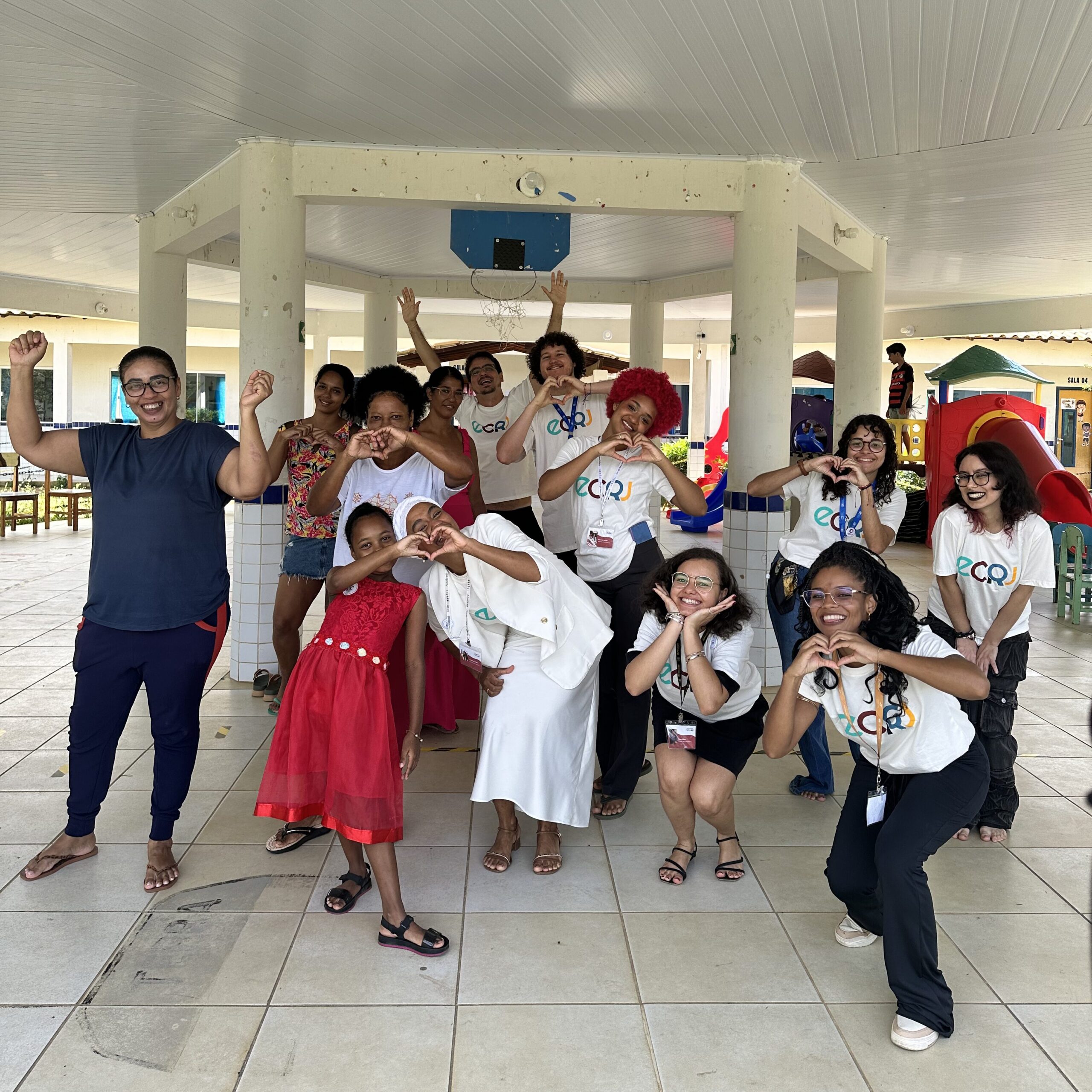 Um grupo de treze pessoas posa alegremente para uma foto em um pátio escolar coberto. No centro, uma mulher em traje branco e uma criança em um vestido vermelho fazem gestos de coração com as mãos. Ao redor delas, outras mulheres e dois homens sorriem.
