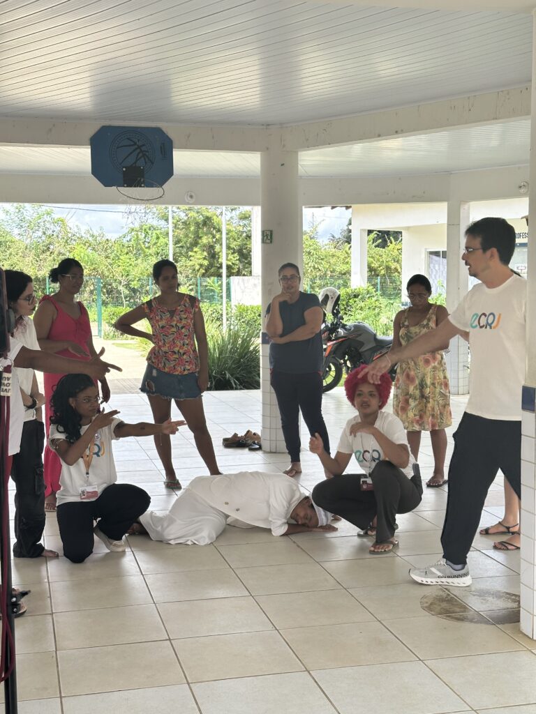 Um grupo de pessoas realiza uma encenação em um pátio coberto. No centro, uma mulher vestida de branco está deitada no chão. Ao lado dela, um homem e duas mulheres com camisetas brancas gesticulam e apontam em sua direção. Ao fundo, cinco mulheres observam a cena em pé. Elas vestem roupas coloridas, como um vestido rosa, uma blusa floral e um vestido amarelo. O ambiente possui pilares brancos, chão de piso claro e vista para um jardim externo.