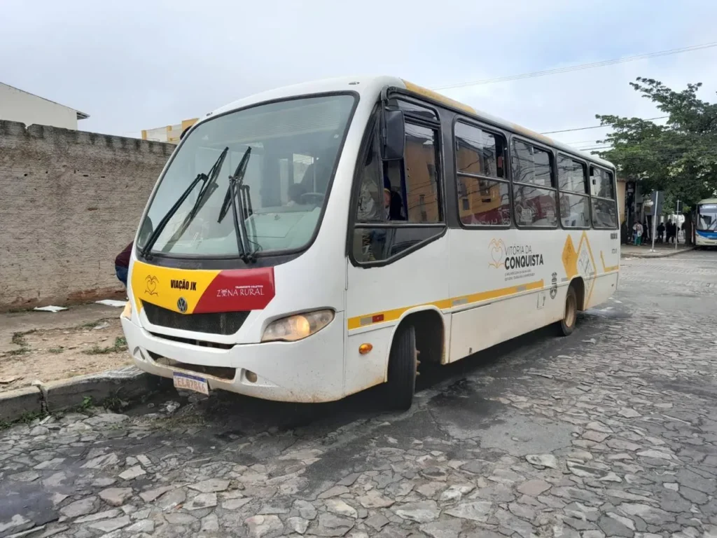 Ônibus branco estacionado em uma rua de paralelepípedos. O veículo possui detalhes em amarelo e vermelho. Na lateral, lê-se "VITÓRIA DA CONQUISTA" acompanhado do brasão do município.