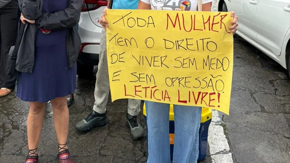 Cartaz amarelo retangular segurado por uma pessoa em uma rua com carros ao fundo. O texto no cartaz diz: TODA MULHER TEM O DIREITO DE VIVER SEM MEDO E SEM OPRESSÃO. LETÍCIA LIVRE!