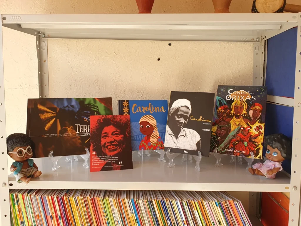 Cinco livros e dois bonecos negros estão expostos em uma prateleira de metal branca. Os livros estão apoiados em suportes transparentes e destacam personalidades e cultura negra. Os títulos e textos visíveis são: "Terreiros", "Carolina", "Contos dos Orixás" e um livro vermelho com o rosto de Angela Davis.