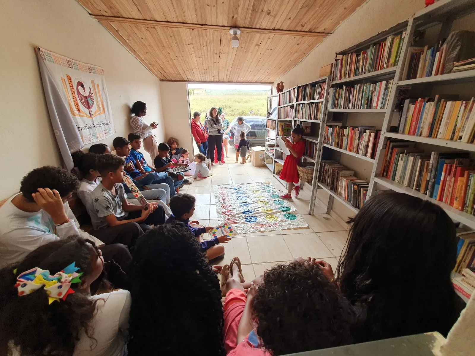 Crianças e adolescentes estão reunidos em uma biblioteca comunitária. No centro, uma menina de vestido vermelho lê um livro em pé. O grupo está sentado no chão e em bancos, atento à leitura. Um tapete de jogo colorido está estendido no piso de cerâmica clara. À direita, estantes metálicas cinzas estão repletas de livros coloridos.