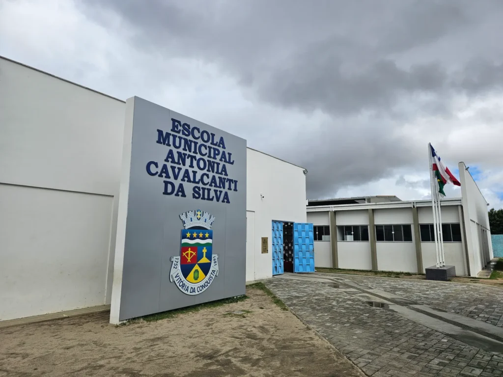Fachada de uma escola pública de alvenaria branca sob um céu nublado. Em destaque, um grande painel cinza traz o nome "ESCOLA MUNICIPAL ANTONIA CAVALCANTI DA SILVA" em letras azuis e, logo abaixo, o brasão do município com o texto "1752 VITÓRIA DA CONQUISTA 1891". À direita do painel, um portão azul de metal está aberto, dando acesso ao pátio interno pavimentado com blocos de concreto. Mais ao fundo, vê-se a lateral das salas de aula com janelas de vidro escuro e, no canto direito, três mastros exibem bandeiras hasteadas.