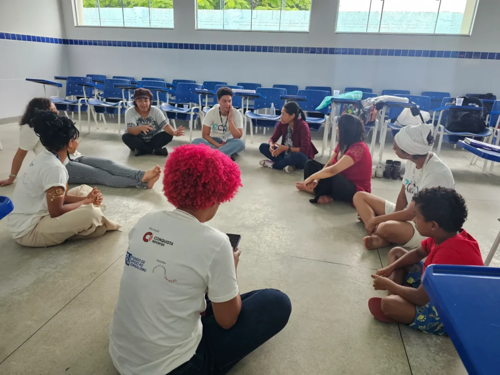 Um grupo de dez pessoas sentadas em círculo no chão de uma sala ampla. Uma pessoa em primeiro plano tem cabelos crespos em tom rosa vibrante e veste uma camiseta branca. Os outros participantes vestem roupas variadas, como camisetas brancas, blusas vermelhas e vinho, e estão descalços ou de chinelos.