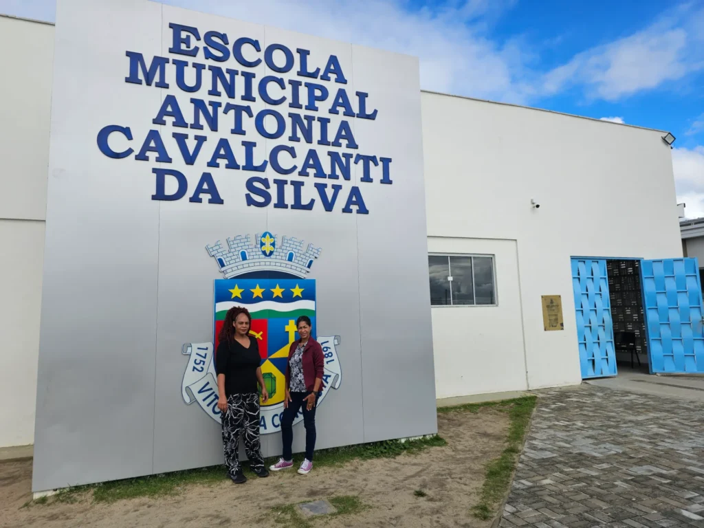 Duas mulheres posam em frente a um grande painel cinza na fachada de uma escola branca sob o céu azul com nuvens. O painel exibe o nome "ESCOLA MUNICIPAL ANTONIA CAVALCANTI DA SILVA" em letras azuis e o brasão do município com a inscrição "1752 VITÓRIA DA CONQUISTA 1891". À esquerda, uma mulher de blusa preta e calça estampada preta e branca sorri discretamente. À direita, a outra mulher veste blazer vinho, blusa florida e calça jeans escura. Ao fundo, um portão metálico azul claro está aberto ao lado de uma área pavimentada.