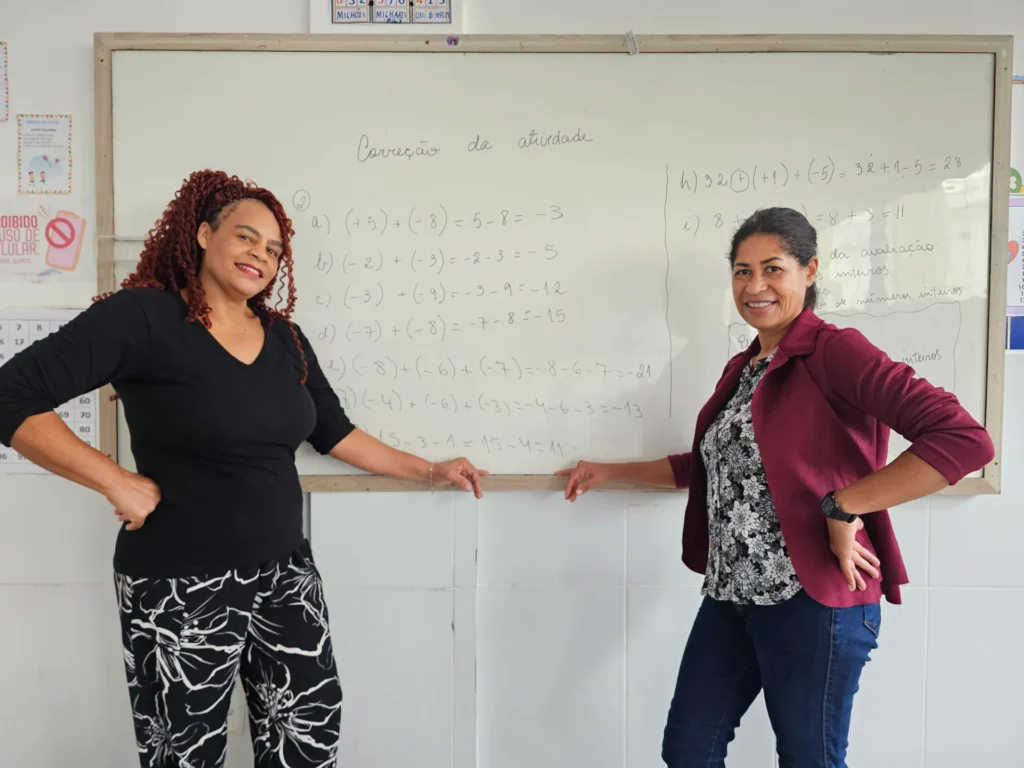 Duas mulheres sorridentes posam lado a lado em frente a um quadro branco de moldura de madeira em uma sala de aula de paredes claras. À esquerda, uma mulher de cabelos cacheados avermelhados, blusa preta e calça estampada preta e branca aponta para o quadro. À direita, a outra mulher veste blazer vinho, blusa florida preta e branca e calça jeans escura, também apontando para o quadro. O quadro traz escrito a caneta o título "Correção da atividade" e uma lista de operações matemáticas.