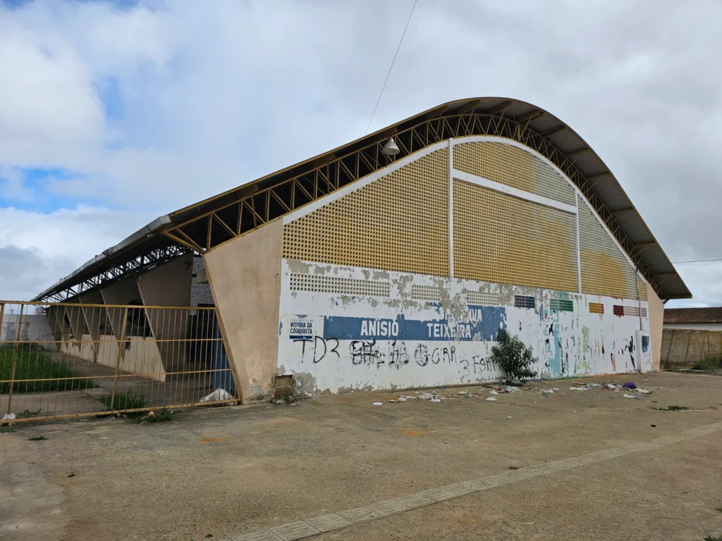 Ginásio grande com teto curvo metálico e paredes em tons de bege e branco, com pintura desgastada e pichações. Na parede frontal aparece o texto “Prefeitura Vitória da Conquista” e “Anísio Teixeira”. O portão lateral está fechado e há lixo espalhado no chão de concreto. O céu está nublado, com algumas áreas azuis aparecendo entre as nuvens.