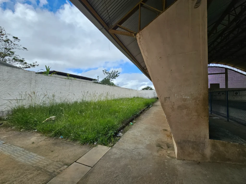 Área externa ao lado de uma quadra coberta, com piso de concreto e parede branca ao fundo. Há mato alto e lixo espalhado perto da parede. À direita, aparece parte da estrutura da quadra, com coluna bege marcada por pichações e grade metálica azul. O céu está nublado, com partes azuis visíveis entre as nuvens.