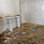 Banheiro com três pias brancas encostadas em paredes de azulejo branco. Uma das pias está quebrada. O chão está coberto de terra, entulho e lixo espalhado, como garrafas plásticas, papéis e pedaços de material de construção. As pias e os azulejos estão sujos e manchados. O ambiente parece abandonado e sem manutenção.