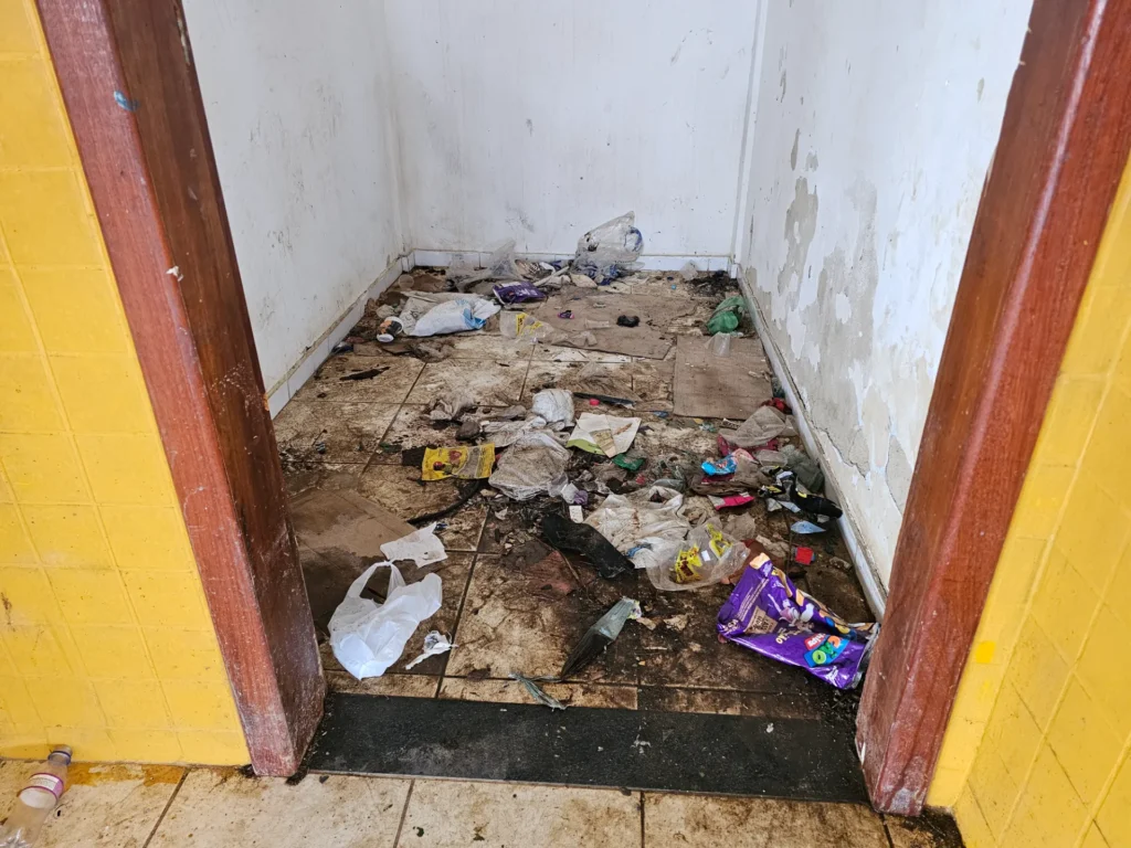 Pequeno cômodo com paredes brancas e entrada com moldura de madeira. O chão de cerâmica está muito sujo, com lixo espalhado, como sacolas plásticas, embalagens, papéis e garrafas. Há manchas escuras no piso e sinais de infiltração e descascamento nas paredes. As paredes externas próximas à porta são amarelas.