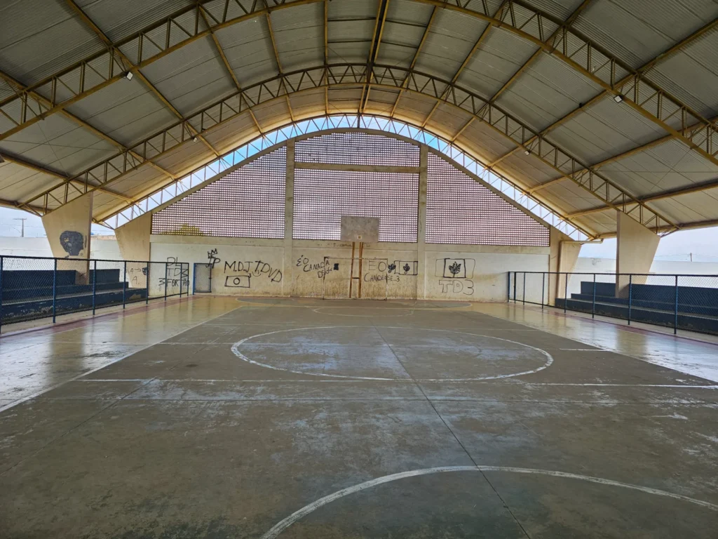 Quadra poliesportiva coberta e vazia. O piso de cimento cinza possui linhas brancas marcadas e uma estrutura de basquete com tabela ao fundo. Uma grande cobertura metálica em arco, com telhas onduladas e vigas amarelas, protege o espaço. Ao fundo, uma parede clara exibe elementos vazados no topo e várias pichações pretas na parte inferior. Nas laterais da quadra, há arquibancadas de cimento escuro protegidas por grades de metal azul.