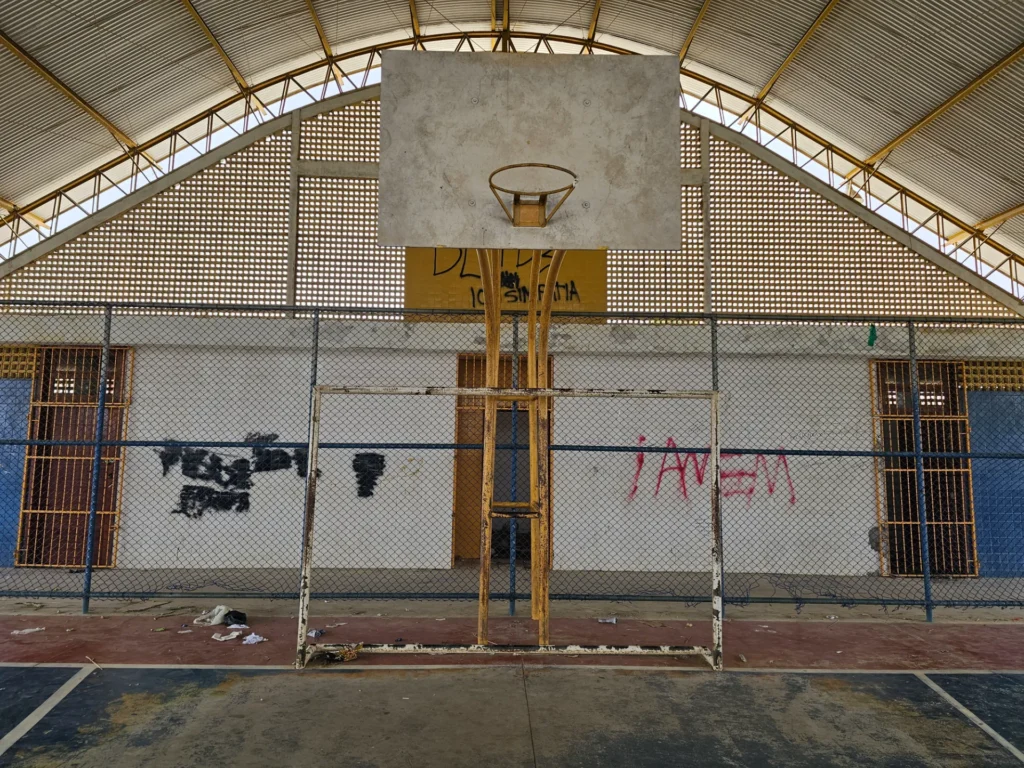 Quadra esportiva coberta com tabela de basquete branca e aro enferrujado no centro. Uma grade metálica separa a quadra da parede ao fundo. As paredes são brancas, com pichações em preto e vermelho. O piso está gasto e há lixo espalhado próximo à grade. O teto é alto, com estrutura metálica e telhas claras. Não há pessoas no local.