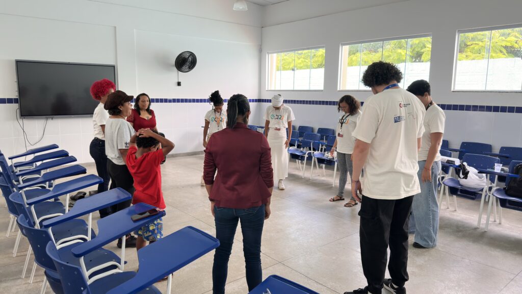 Um grupo de pessoas forma um círculo no centro de uma sala de aula ampla e iluminada. Elas estão de pé, algumas com cabeças levemente inclinadas. Ao redor, há diversas cadeiras azuis organizadas junto às paredes brancas.