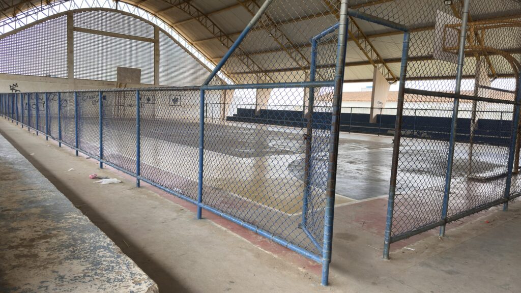 Uma cerca de tela metálica azul e cinza cruza toda a extensão separando o corredor de acesso da quadra poliesportiva interna. A quadra possui piso de cimento cinza polido, cercada por arquibancadas escuras ao fundo. Uma grande cobertura metálica com vigas amarelas protege todo o espaço. À esquerda, em primeiro plano, destaca-se uma mureta baixa de cimento áspero e desgastado. Na parede branca ao fundo, há elementos vazados na parte superior e pequenas pichações pretas ilegíveis na parte inferior, além de marcas de uma tabela de basquete.