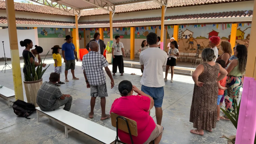Grupo de pessoas participa de uma atividade em roda em um pátio coberto com colunas amarelas e piso de cimento. Algumas pessoas estão em pé e outras sentadas em bancos brancos. Jovens com camisetas brancas com a sigla “ECRJ” conduzem a atividade no centro da roda. Ao fundo, as paredes têm desenhos coloridos, bandeirinhas decorativas e cadeiras alinhadas.