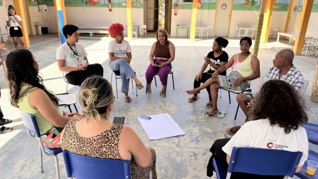 Grupo de pessoas está sentado em roda em cadeiras azuis em um pátio coberto com colunas amarelas. Jovens com camisetas brancas com a sigla “ECRJ” participam da conversa ao lado de homens e mulheres adultos. No chão, há uma folha branca grande e um celular. Ao fundo, aparecem mesas, cadeiras brancas e paredes com desenhos coloridos. Na camiseta de uma participante aparece o texto “Conquista Repórter”.