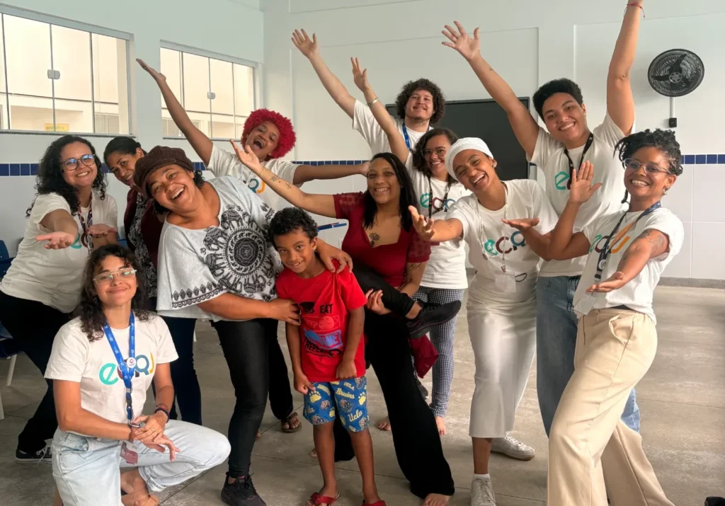 Um grupo diverso de cerca de doze pessoas sorri e posa com braços abertos em uma sala ampla de paredes brancas. A maioria veste camisetas brancas com o logotipo colorido "ecrj". Ao centro, uma criança em uma camiseta vermelha é abraçada por adultos.