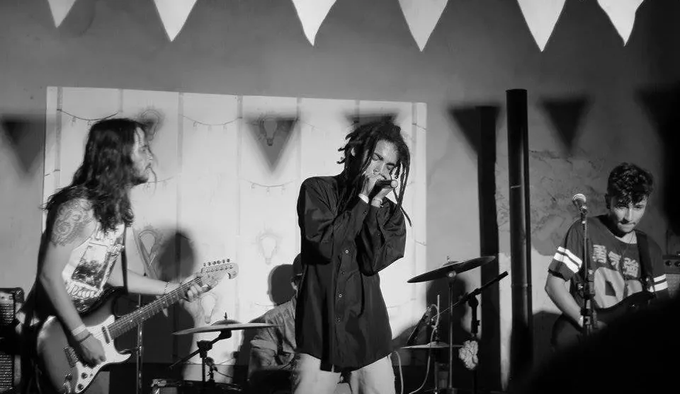 Uma banda de rock se apresenta em um palco iluminado, em uma fotografia em preto e branco. No centro, o vocalista, um homem negro com cabelos de dreadlocks longos, veste camisa preta e calça clara, e canta segurando o microfone próximo à boca com as duas mãos e os olhos fechados. À esquerda, um guitarrista de cabelos longos e tatuagem no braço toca uma guitarra elétrica. À direita, outro músico de camiseta escura toca um instrumento. Ao fundo, um baterista está posicionado atrás dos pratos da bateria.