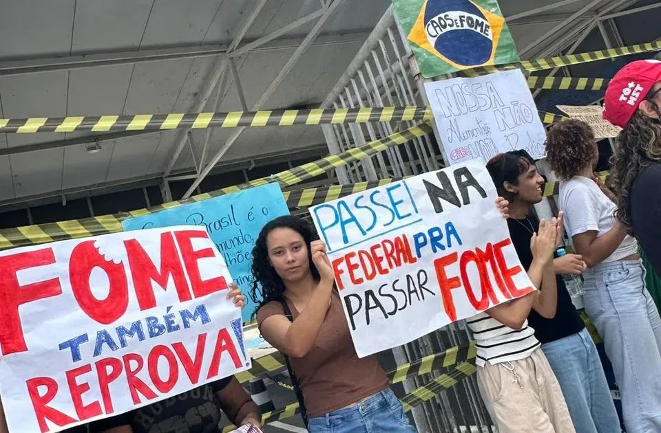 Estudantes protestam em frente a um prédio interditado. Em primeiro plano, uma jovem de cabelos cacheados e blusa marrom segura um cartaz com letras grandes. O cartaz diz: "PASSEI NA FEDERAL PRA PASSAR FOME".