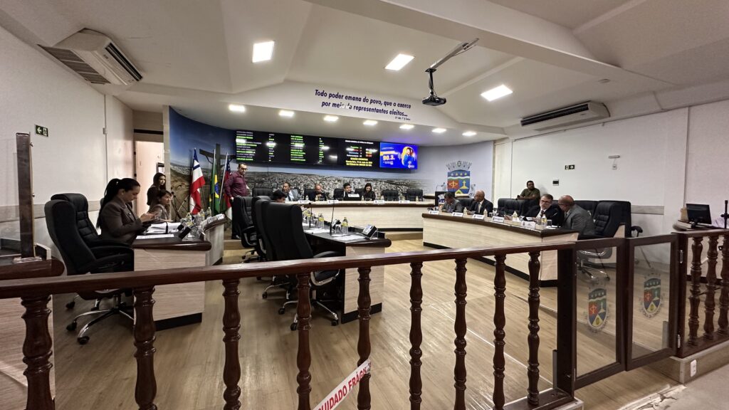 Plenário de câmara municipal com vereadores sentados em mesas em formato semicircular. O ambiente tem piso amadeirado, cadeiras pretas, paredes claras e iluminação branca no teto. Ao fundo, há bandeiras e painéis eletrônicos com informações da sessão. Algumas pessoas conversam enquanto outras usam celular ou computador. Na parede superior está escrito: “Todo poder emana do povo, que o exerce por meio de representantes eleitos...” e abaixo “C. F. 1º”.