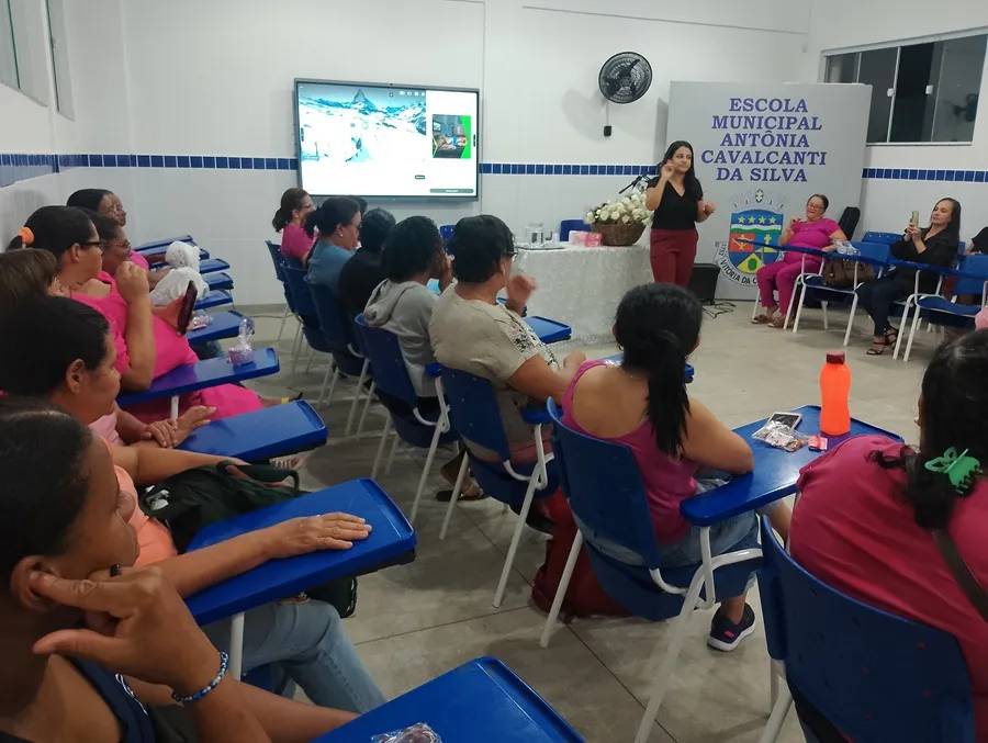 Mulheres adultas assistem a uma palestra sentadas em carteiras universitárias azuis dentro de uma sala de aula de paredes brancas e detalhes em azulejos azuis. À frente do grupo, uma palestrante de blusa preta e calça escura fala de pé ao lado de uma mesa com toalha branca e arranjo de flores. Ao fundo, um painel cinza exibe o texto "ESCOLA MUNICIPAL ANTÔNIA CAVALCANTI DA SILVA" junto ao brasão municipal, onde se lê parcialmente "VITÓRIA DA CONQUISTA". Na parede ao fundo, uma tela digital ligada exibe uma paisagem nevada e uma mulher sentada em outra cadeira grava a apresentação com um celular.