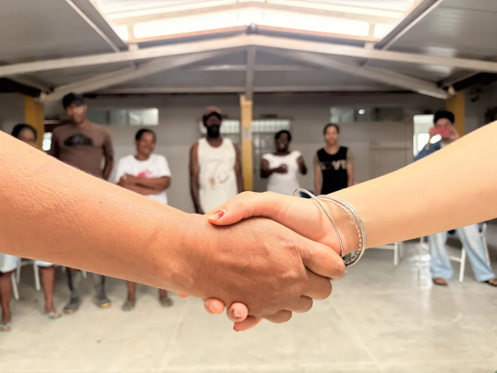 Duas pessoas apertam as mãos no centro da cena, em primeiro plano. Uma mão tem pulseiras prateadas e unhas pintadas. Ao fundo, desfocado, há um grupo de pessoas em pé dentro de um espaço coberto, com estrutura metálica branca e piso claro. Algumas pessoas usam roupas em tons escuros e claros.