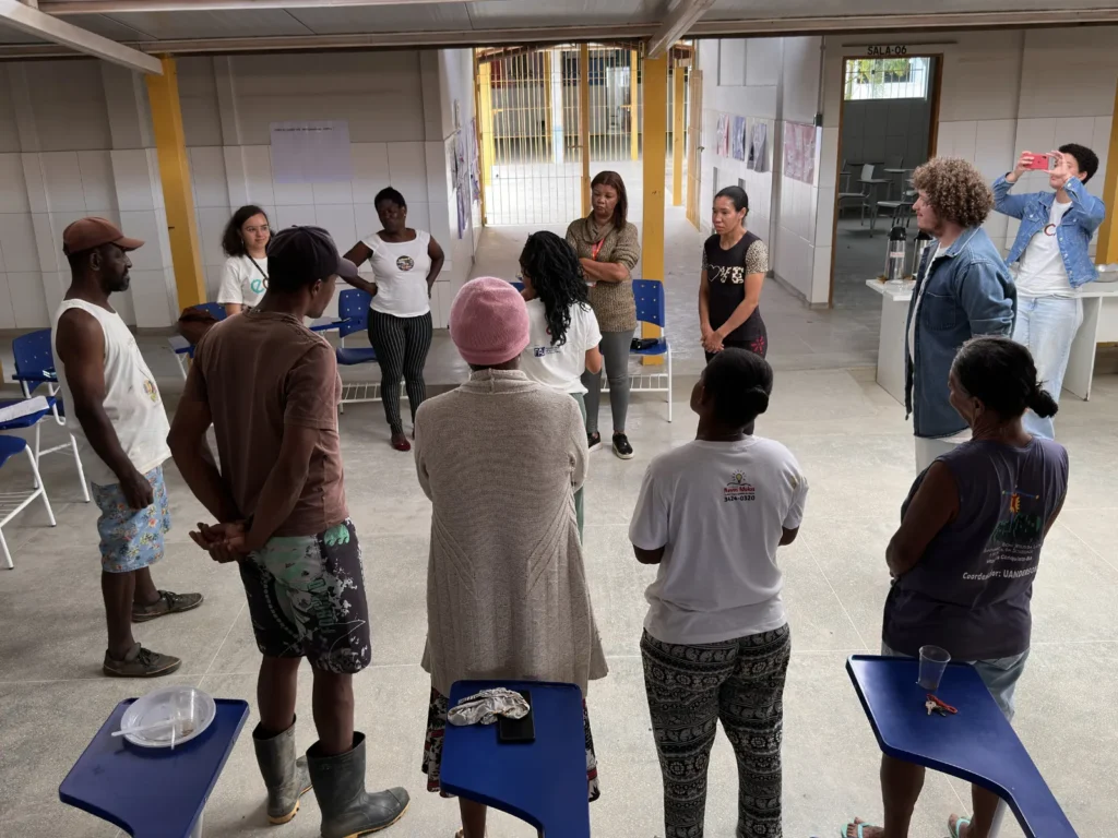 Grupo de pessoas forma um círculo em uma sala ampla com piso claro, paredes brancas e colunas amarelas. Algumas pessoas estão em pé à frente de cadeiras azuis, ouvindo uma mulher no centro do grupo. À direita, uma pessoa tira foto com o celular. Há cartazes nas paredes e uma porta ao fundo com o texto “SALA-06”.