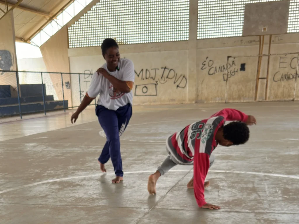 Duas pessoas jogam capoeira em uma quadra coberta de piso cinza. Uma mulher sorri enquanto avança descalça, usando camiseta branca e calça azul. À frente dela, uma criança com blusa vermelha, preta e branca apoia uma mão no chão durante o movimento. Ao fundo, há paredes claras com pichações e uma estrutura metálica no teto.