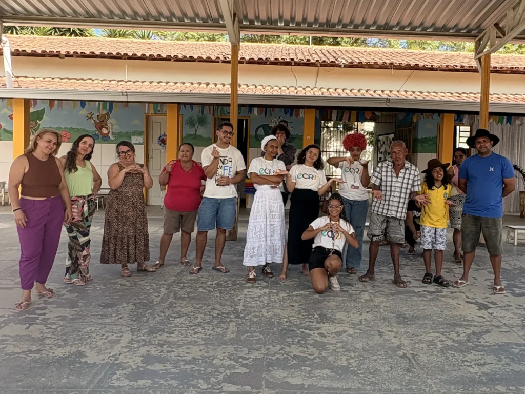 Grupo de pessoas posa em um espaço coberto com piso de cimento e colunas amarelas. Ao centro, jovens e adultos fazem gestos com as mãos e sorriem para a câmera. Algumas pessoas usam camisetas brancas com a sigla “ECRJ”. Ao fundo, há paredes com desenhos coloridos, bandeirinhas e decorativas.