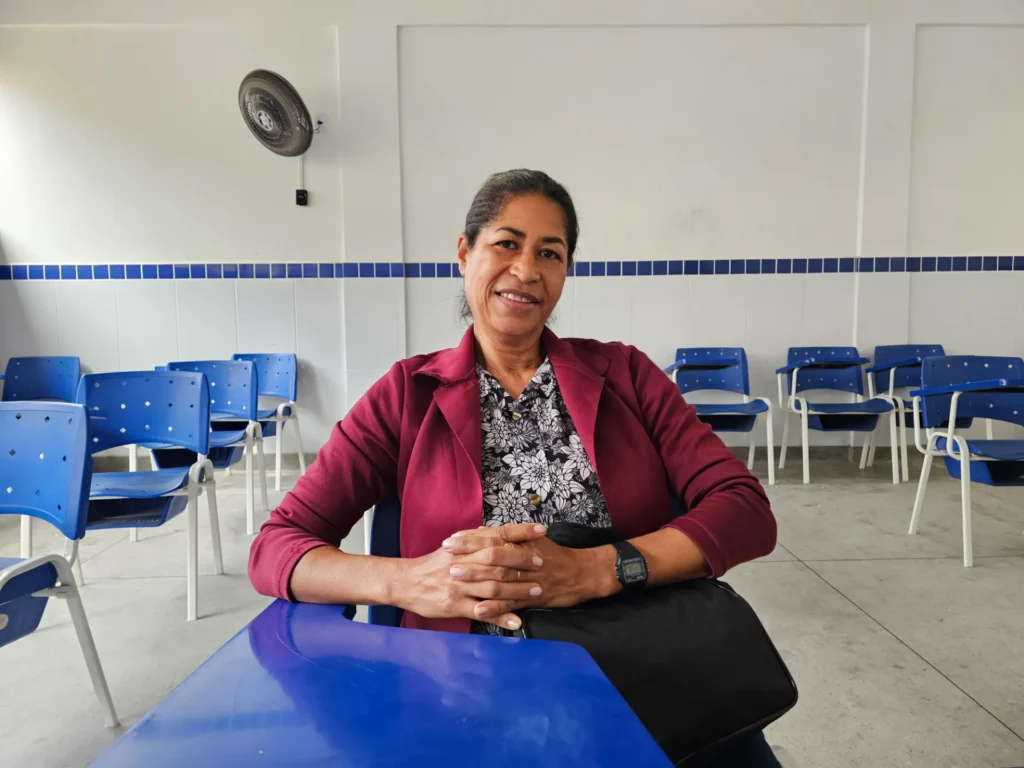 Uma mulher sorridente com cabelo preto preso em um coque baixo e blazer cor de vinho sobre uma blusa florida preta e branca senta-se atrás de uma carteira azul com as mãos entrelaçadas sobre uma bolsa preta. Ela está sentada em uma sala de aula vazia com fileiras de cadeiras azuis de assento e encosto correspondentes organizadas em fileiras ordenadas atrás dela. As paredes da sala de aula são brancas com uma faixa de azulejos azuis. Um ventilador de teto branco está pendurado na parede acima e atrás dela.