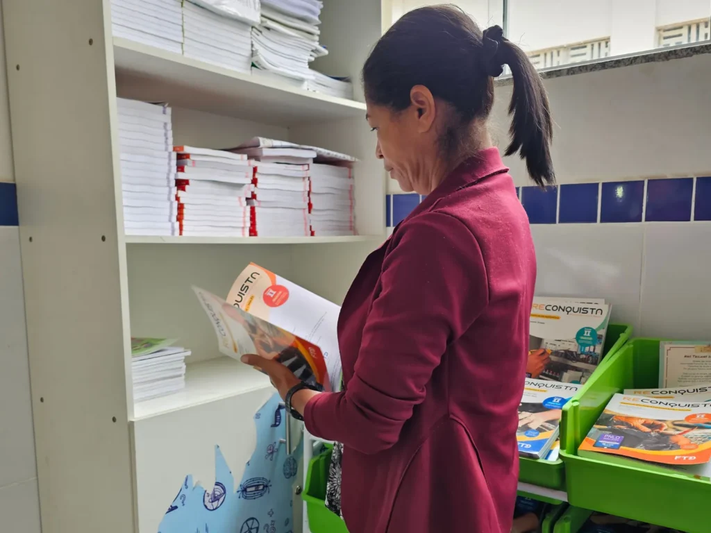 Uma mulher de perfil, com cabelo preto preso em um rabo de cavalo e blazer vinho, folheia um livro didático de capa branca e detalhes em laranja. Ela está em pé diante de uma estante branca repleta de pilhas de livros. Ao seu lado direito, caixas plásticas verdes guardam mais apostilas. Na capa de uma das apostilas abertas e nas que estão empilhadas nas caixas, lê-se a palavra "RECONQUISTA". O ambiente possui paredes brancas com uma faixa de azulejos azuis escuros.