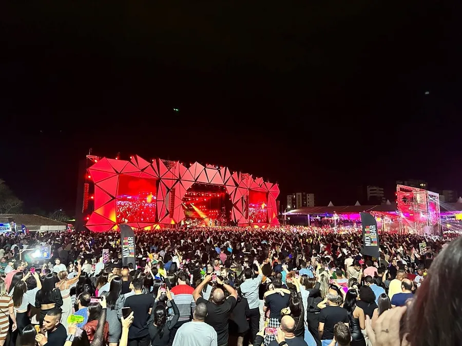 Multidão lota um festival de música ao ar livre durante a noite. Ao fundo, um grande palco iluminado de vermelho possui estrutura geométrica com triângulos e dois telões laterais. Centenas de pessoas estão de costas, muitas delas erguendo celulares para registrar o show. À direita, uma bandeira preta exibe o logotipo colorido MIX BAR. O céu está totalmente escuro e prédios aparecem de forma discreta no horizonte.