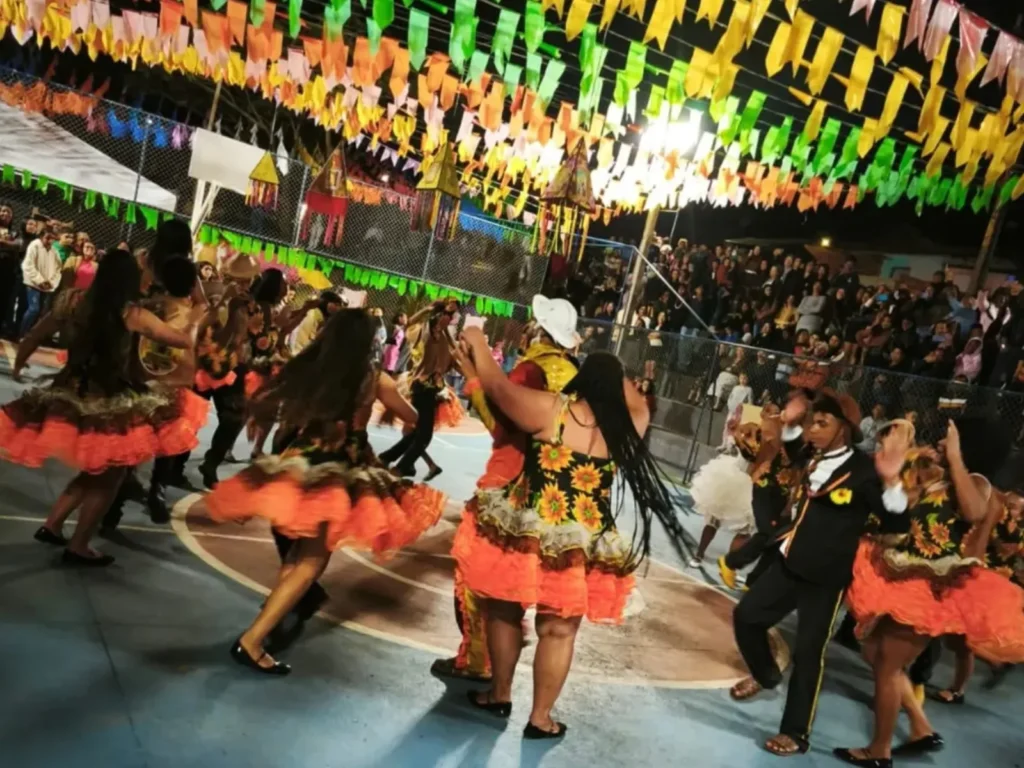 Pessoas dançam quadrilha em uma quadra esportiva durante uma festa junina à noite. Os dançarinos usam trajes típicos coloridos, com destaque para vestidos rodados de chita com estampas de girassol e babados laranjas. No teto, fileiras de bandeirinhas coloridas em tons de amarelo, verde e laranja cruzam todo o espaço. Ao fundo, uma rede de proteção separa a quadra do público que assiste à apresentação. Lanternas de papel decorativas pendem do teto junto às bandeiras.