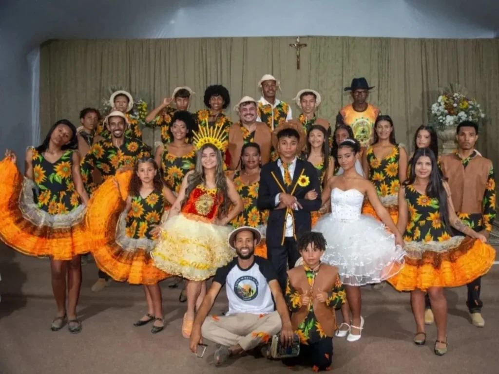 Vinte e três pessoas formam um grupo de quadrilha. No centro, uma noiva com vestido branco rodado dá o braço a um noivo de terno e gravata de girassol, ao lado de uma rainha com vestido laranja e coroa. Outras mulheres usam vestidos de chita com estampa de girassol e babados laranjas, amarelos e verdes, que elas seguram abertos. Os homens vestem camisas de girassol e chapéus brancos. Duas crianças e um homem sentado com um livro completam o grupo na frente. O cenário é um auditório com cortinas marrons e dois arranjos florais amarelos e laranjas em pedestais ao fundo.