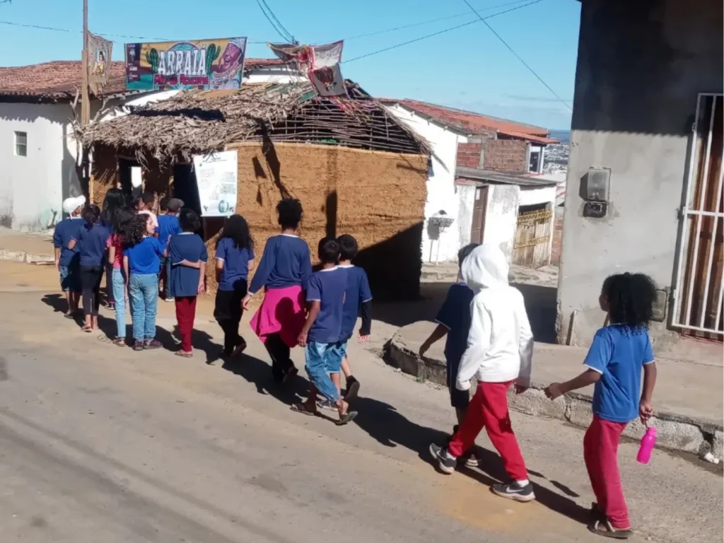 Grupo de crianças caminham em fila indiana por uma rua de asfalto em direção a uma casa de taipa. A maioria veste uniformes escolares azuis. A casa ao fundo é feita de barro com telhado de palha seca. Acima da entrada, um banner colorido exibe o texto "ARRAIÁ Flor de Açucena". O dia está ensolarado com céu azul limpo, projetando sombras nítidas no chão. Outras casas simples compõem o cenário ao redor.