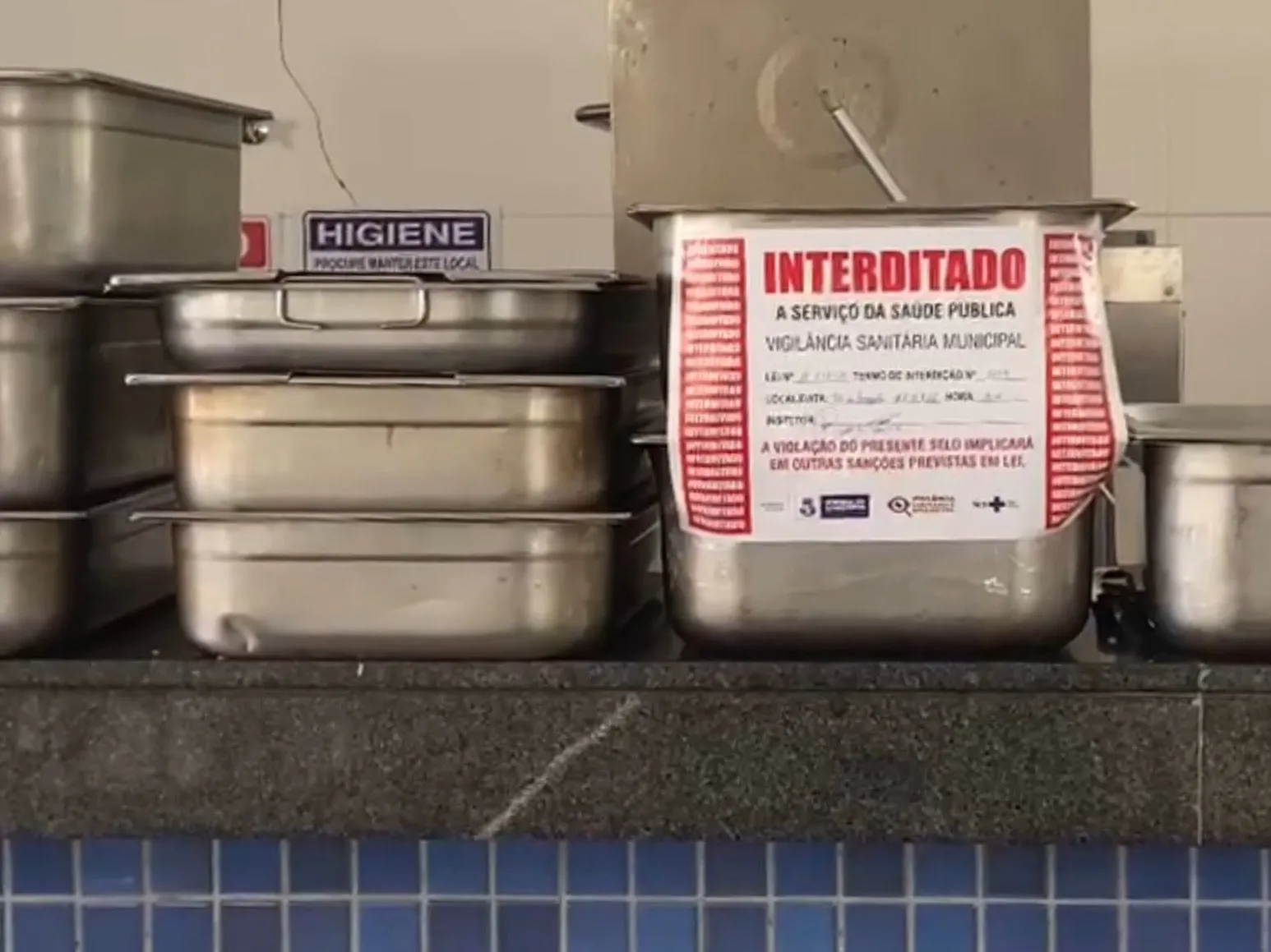 Recipiente de metal em uma cozinha industrial com um adesivo de interdição colado na frente. O adesivo é branco com bordas vermelhas e traz a palavra "INTERDITADO" em destaque, seguida pelos textos: "A SERVIÇO DA SAÚDE PÚBLICA", "VIGILÂNCIA SANITÁRIA MUNICIPAL" e um aviso de que a violação do selo implicará em sanções legais. Ao lado e atrás, há outros recipientes de aço inox empilhados sobre um balcão de granito escuro. No fundo, uma parede clara exibe uma placa azul e branca com a palavra "HIGIENE" e a frase "Procure manter este local". A base do balcão é revestida por azulejos azuis pequenos.