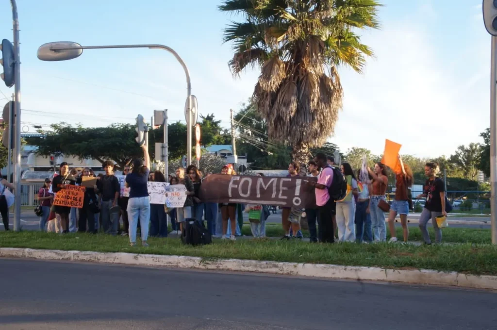Grupo de estudantes protesta em um canteiro ao lado de uma avenida, segurando cartazes e uma faixa marrom com a palavra “FOME” em letras brancas. Um dos cartazes diz: “RU também distribui dignidade e permanência”. Os estudantes usam roupas casuais e mochilas. Ao fundo há um coqueiro alto, postes, semáforos e árvores. A cena acontece no fim da tarde, com luz natural e céu claro.