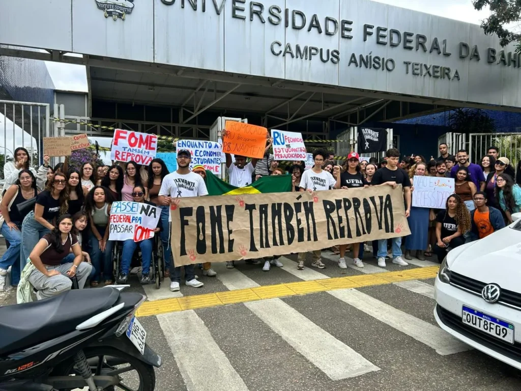 Grupo de estudantes protesta em frente à entrada da Universidade Federal da Bahia, Campus Anísio Teixeira. No centro, várias pessoas seguram uma faixa bege com a frase “FOME TAMBÉM REPROVA”. Outros cartazes dizem: “Fome também reprova”, “RU fechado = estudantes prejudicados”, “Passei na Federal para passar fome”, “Promoção RU dos sonhos R$ não tem” e “Enquanto a economia, estudantes com fome”. Há uma bandeira do Brasil atrás da faixa principal. Os estudantes usam roupas casuais e estão reunidos sobre uma faixa de pedestres. Ao fundo aparece a fachada da universidade.